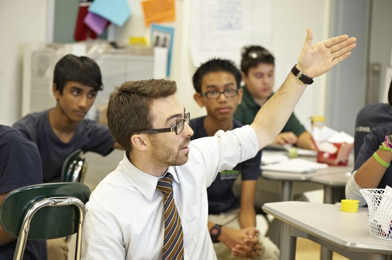 The image shows a classroom setting with a teacher and students. The teacher, a young man with glasses and a tie, is gesturing with his hand raised, possibly asking a question or calling on a student. Several students are visible, some looking at the teacher. The classroom appears to be bright and well-lit, with desks and other school-related items in the background.